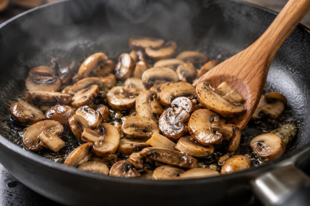 Golden brown sliced mushrooms sauteing in a skillet with wooden spoon
